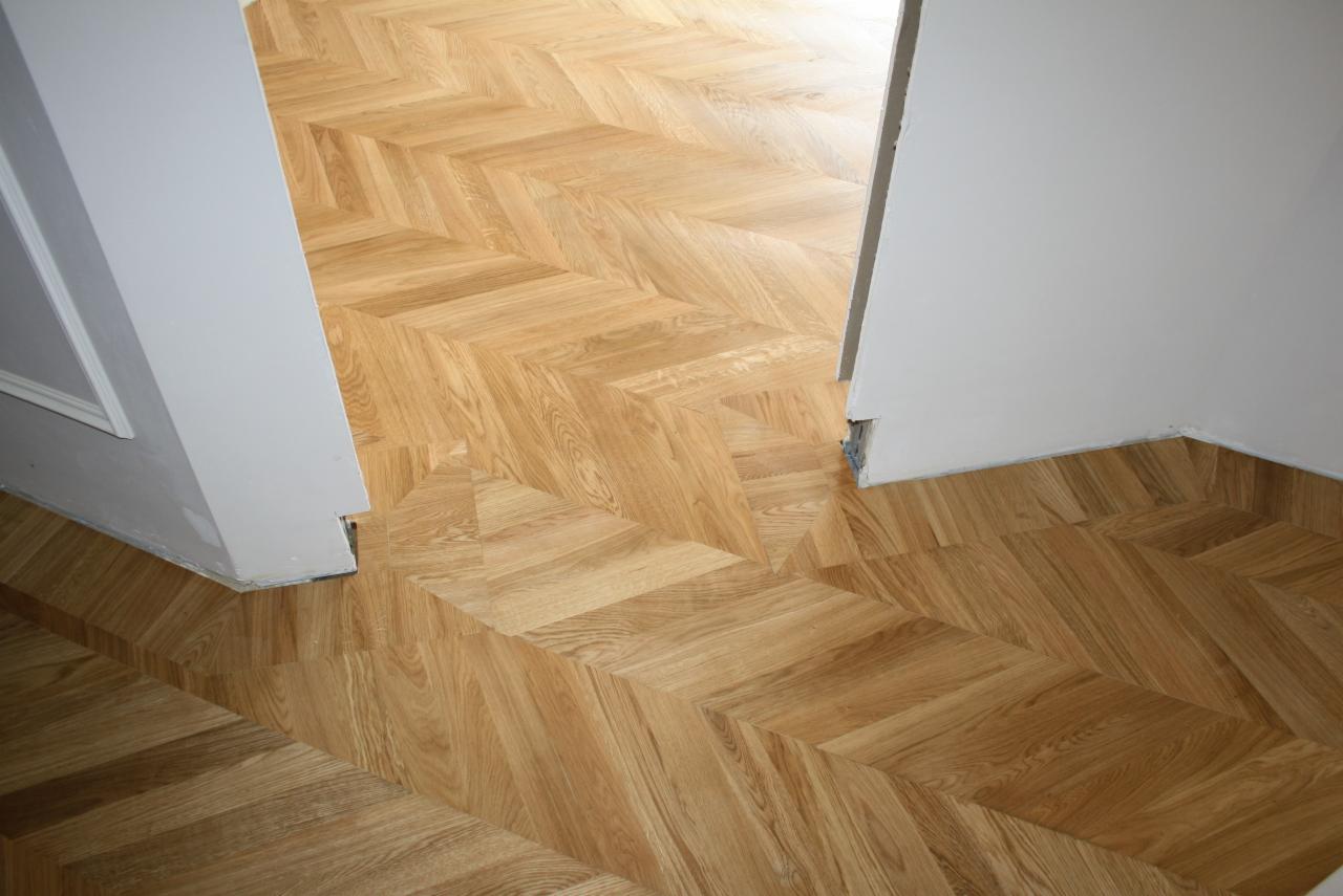 Ułożony parkiet dębowy w jodełkę francuską, widoczne niedoskonałości przy łączeniu z ościeżnicą, jasne ściany.