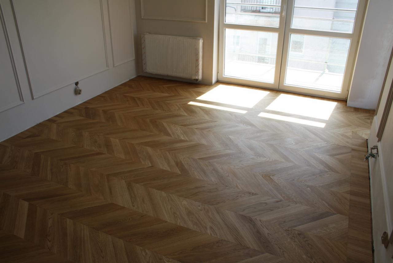 Ułożony parkiet w jodełkę w jasnym pomieszczeniu z oknem i białymi ścianami z dekoracyjnymi listwami, widoczne promienie słoneczne.