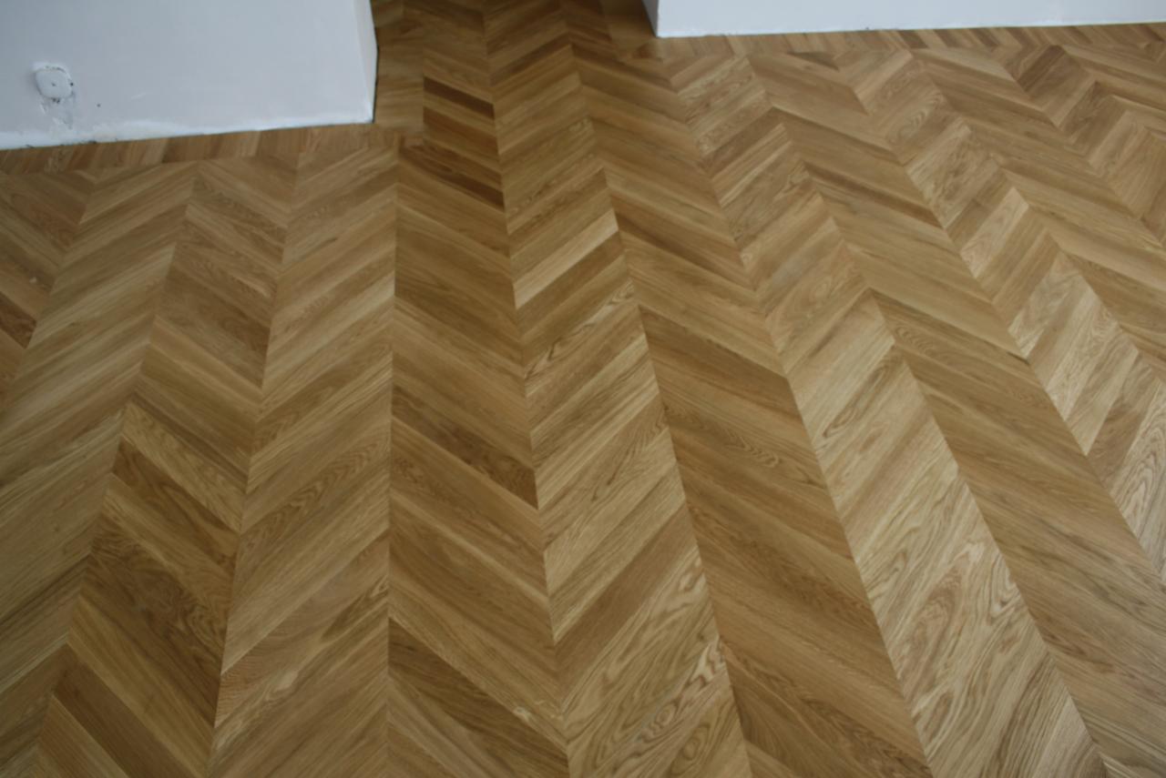Parkiet ułożony w jodełkę z jasnego drewna, świeżo po cyklinowaniu, z widocznymi śladami po szpachlowaniu przy ścianie.