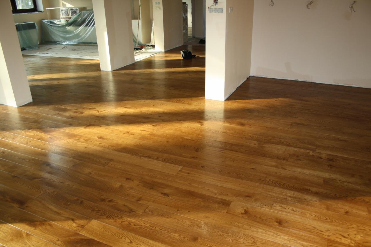 Wykończona podłoga z parkietu w jasnym odcieniu drewna, z widocznymi promieniami słońca padającymi na powierzchnię, w tle przygotowane pomieszczenie do dalszych prac wykończeniowych.