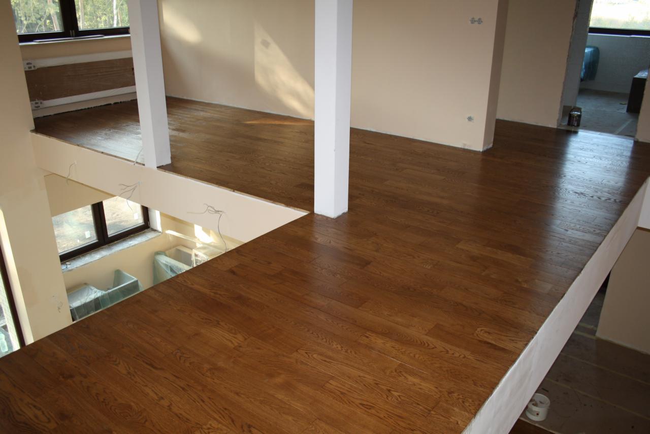 Świeżo położony, lakierowany parkiet w dwupoziomowym wnętrzu w Luboniu, z widocznymi fragmentami instalacji elektrycznej i przygotowaniami do dalszych prac wykończeniowych.