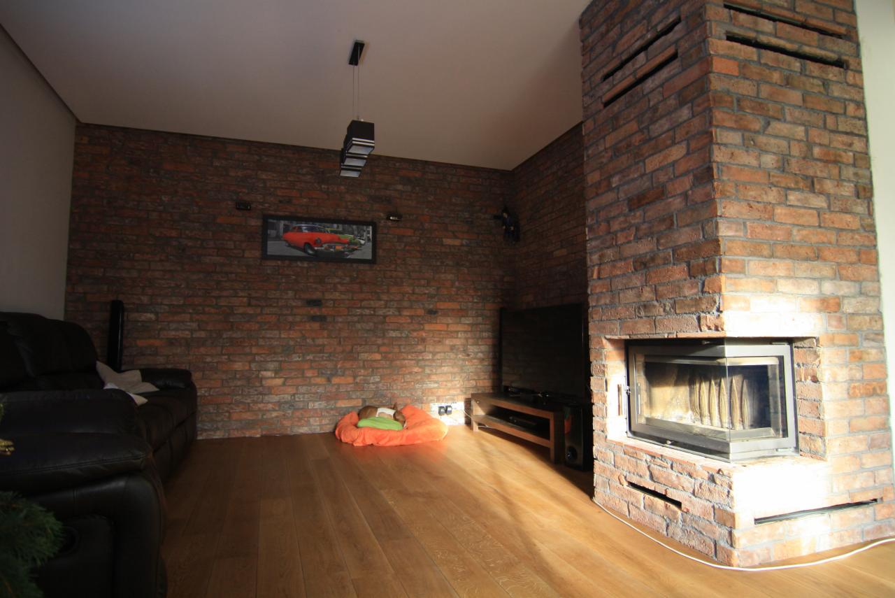 Salon z ceglaną ścianą, kominkiem z cegły, drewnianą podłogą, czarną skórzaną sofą i poduszką dla psa na podłodze.