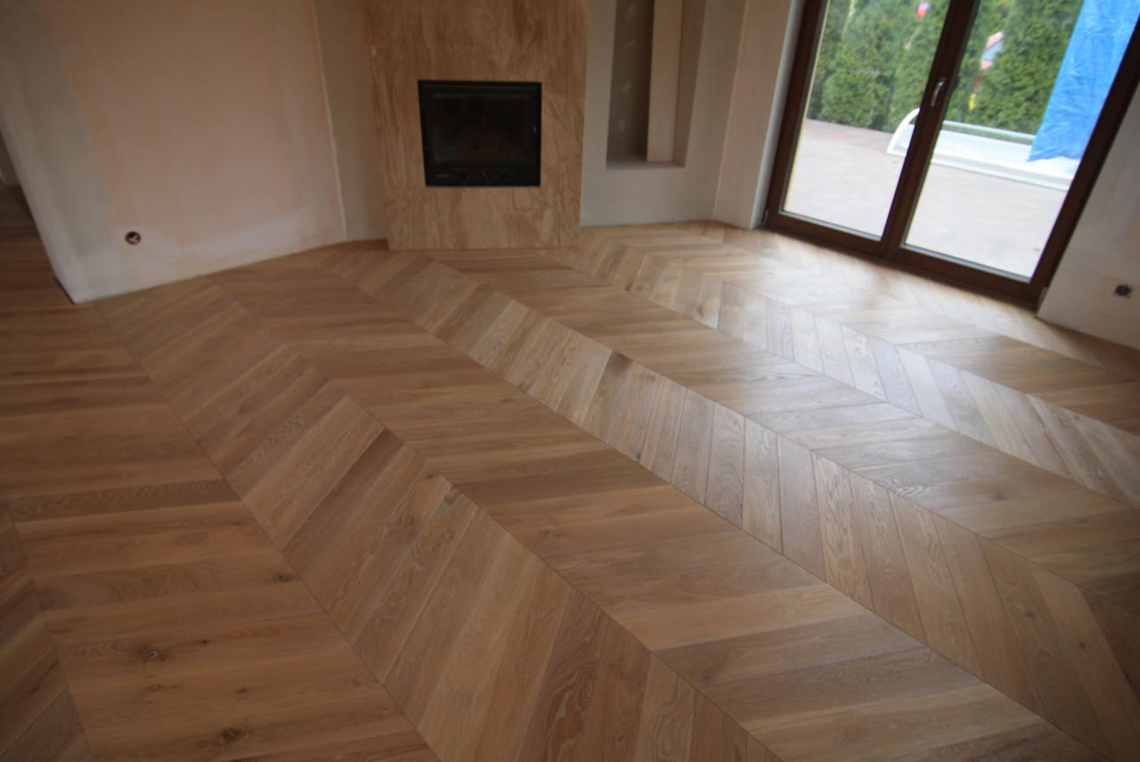 Jasnobrązowa podłoga ułożona w jodełkę francuską, widoczny kominek z marmurowym obramowaniem i duże okno z widokiem na zieleń na zewnątrz.