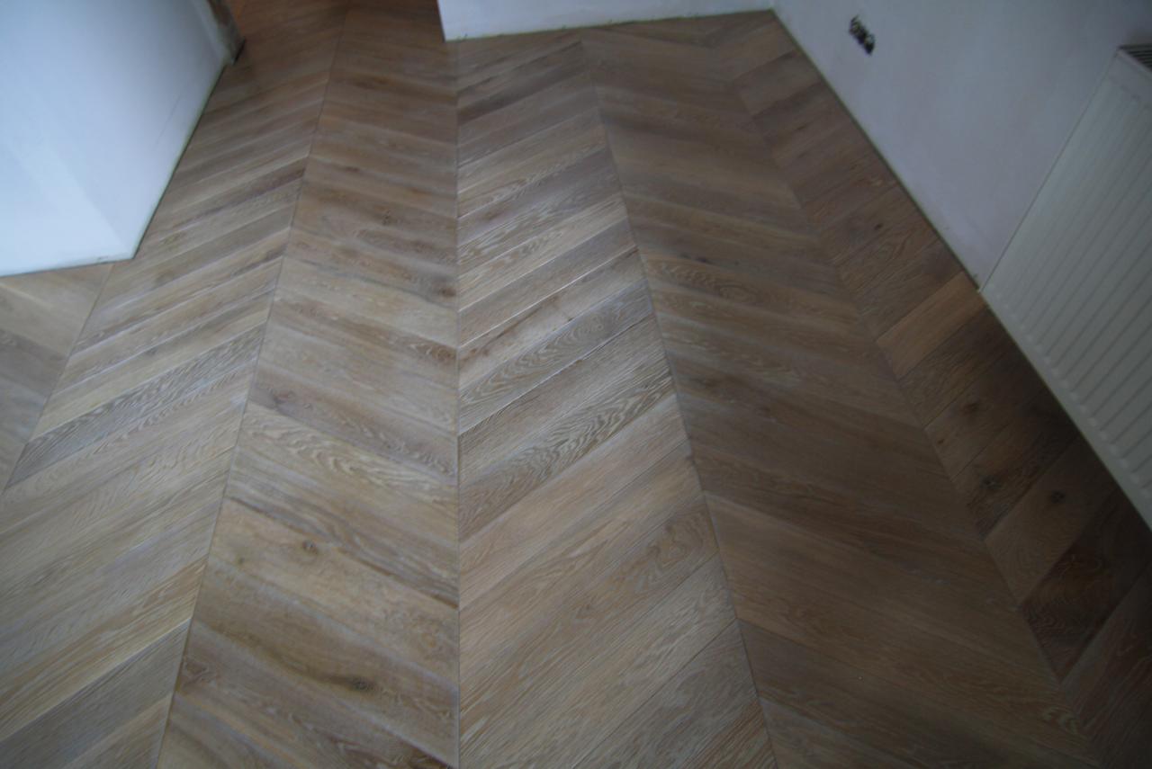 Ułożony parkiet w jodełkę, jasne drewno, widoczne ściany i grzejnik, instalacja elektryczna bez osprzętu.