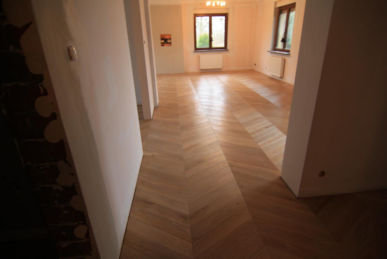 Ułożony parkiet w jodełkę w jasnym, przestronnym pokoju z oknami i grzejnikami, widok zza progu przez otwarte drzwi.