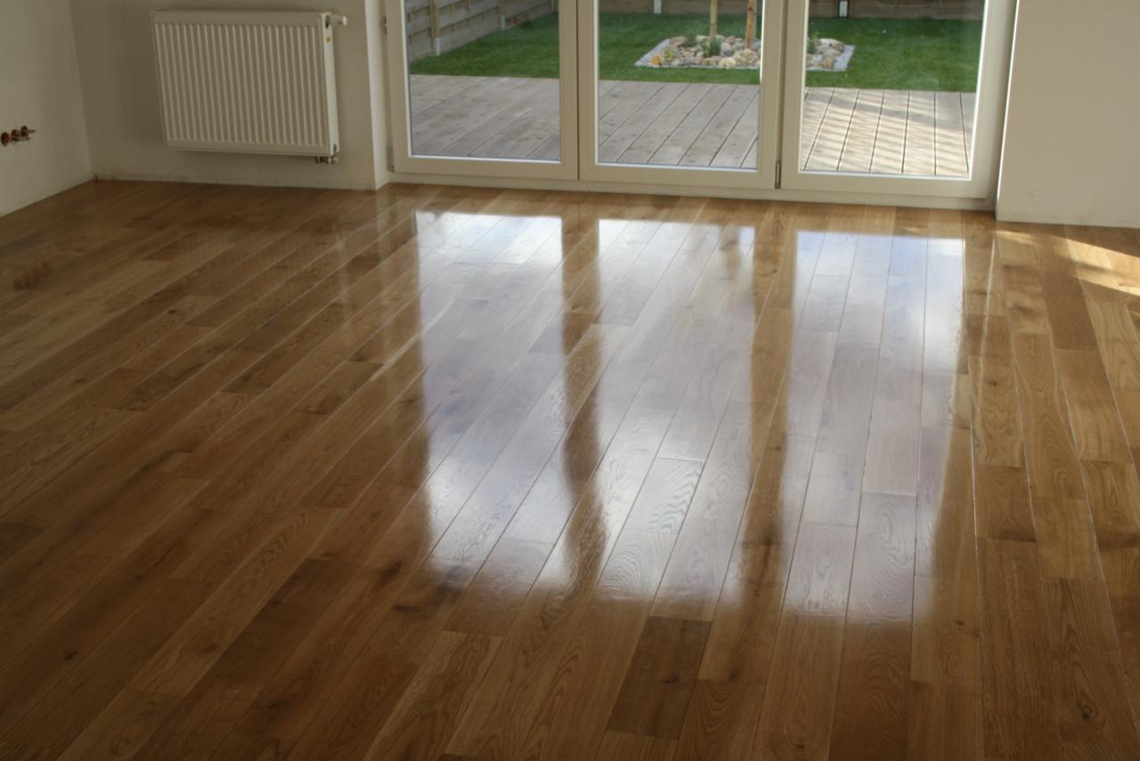 Lśniąca, świeżo polakierowana drewniana podłoga parkietowa w jasnym odcieniu, ułożona w regularny wzór, z widocznymi refleksami światła słonecznego wpadającego przez duże okno balkonowe z widokiem...