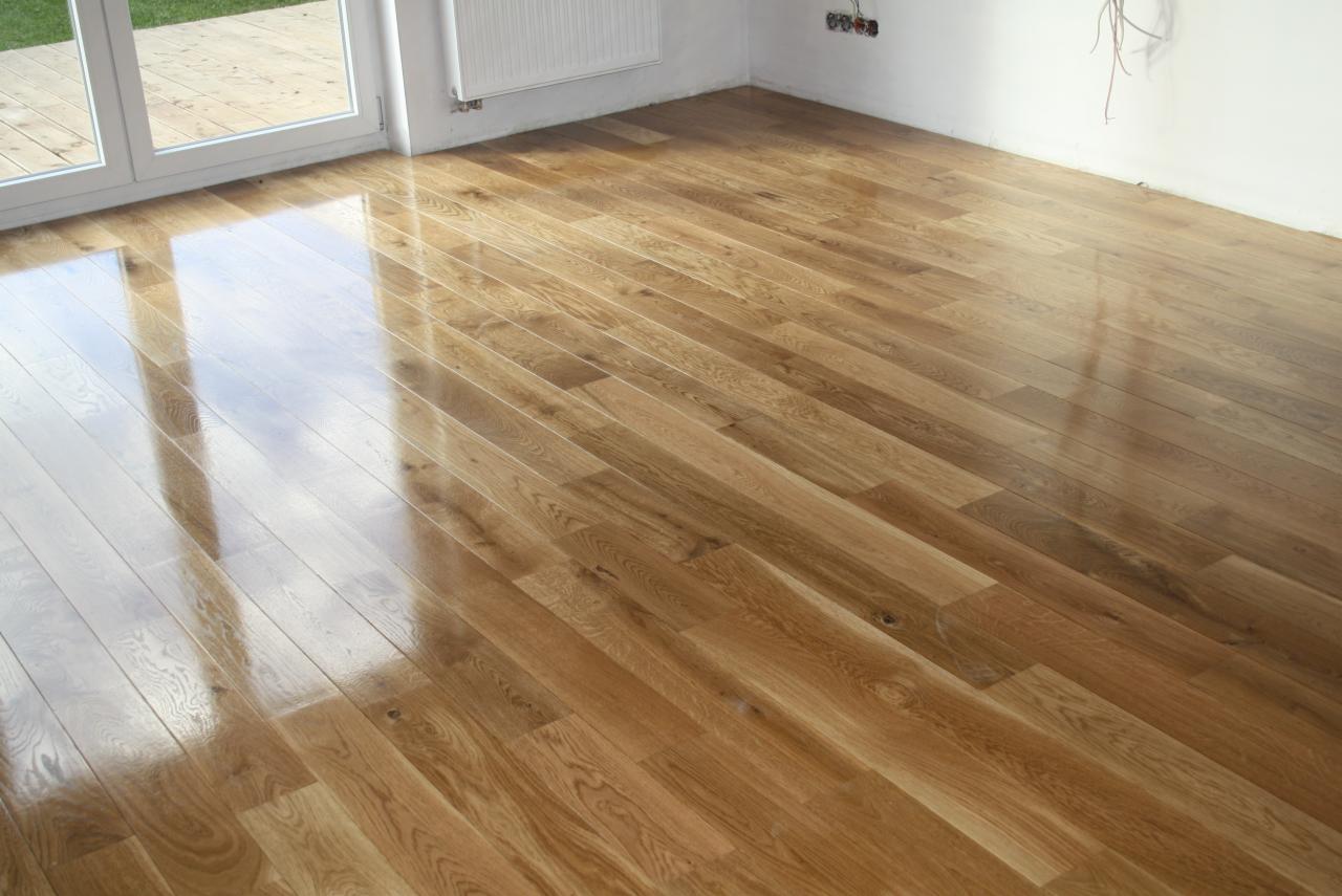 Błyszcząca, świeżo położona parkietowa podłoga z widocznymi słojami drewna, odbijająca światło z okna balkonowego, w rogu białej ściany widoczne gniazdka elektryczne i wystające kable.