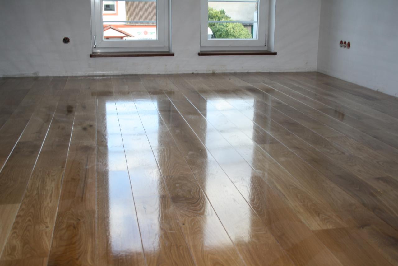 Błyszcząca, świeżo polakierowana podłoga z desek w pustym pokoju z dwoma oknami; widoczne refleksy światła odbijające się od powierzchni drewna.