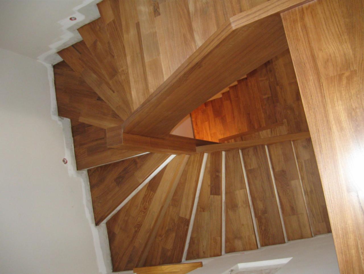 Widok z góry na drewniane schody spiralne z jasnego drewna, z widoczną konstrukcją i wykończeniem, oświetlone od dołu.