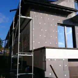 Dom w trakcie ocieplania, widoczne szare płyty styropianowe z białymi kołkami, rusztowanie, okna z odbiciem nieba, w tle zieleń.