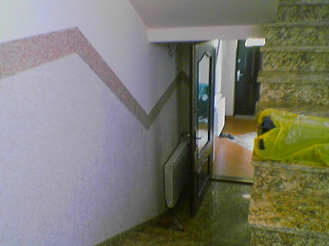 Klatka schodowa z marmurowymi schodami i ścianą z dekoracyjnym, zygzakowatym pasem. Widoczne drzwi wejściowe do mieszkania i grzejnik. Żółta torba leży na schodach.