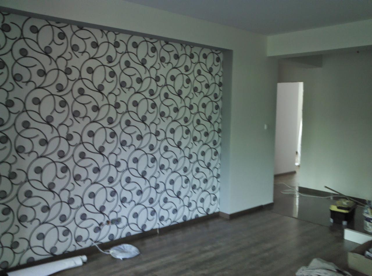 Ściana z wzorzystą tapetą w pokoju, widoczne niedociągnięcia przy listwie przypodłogowej i resztki materiałów na podłodze.