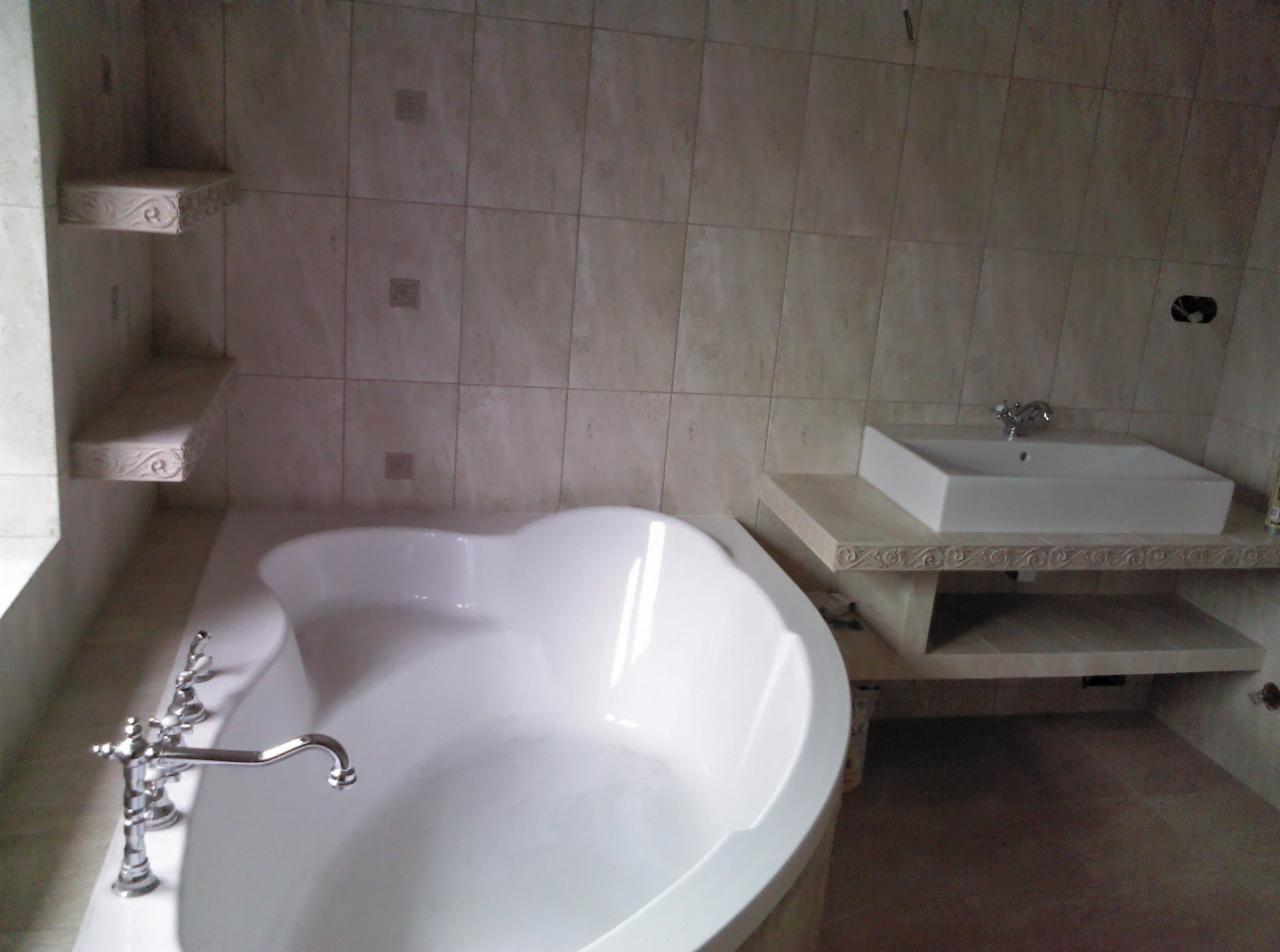 Wykończona łazienka z beżowymi płytkami, owalną wanną z chromowaną armaturą i prostokątną umywalką na zdobionej półce.