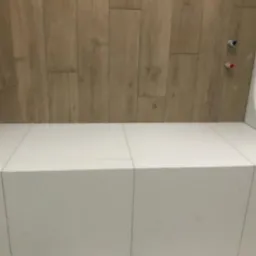 Wykończona obudowa wanny z białych płytek ceramicznych, z drewnopodobną okładziną ściany w tle i widocznymi zaworami.