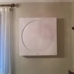 Biały, kwadratowy klimatyzator LG zamontowany na ścianie w pokoju, widoczny fragment firanki i drewniana listwa pod sufitem.