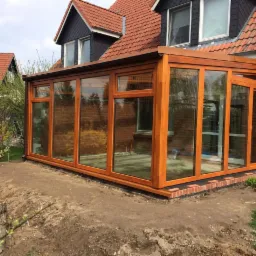 Zewnętrzny widok dobudowanej oranżerii z drewnianą ramą okienną i szklanymi panelami, przylegającej do ceglanego domu z czerwonym dachem. Ziemia wokół przygotowana pod ogród.