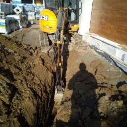 Żółta mini koparka JCB wykonuje wąski wykop w ziemi, cień operatora widoczny na gruncie, w tle materiały budowlane i fragment budynku z drewnianą elewacją.