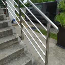 Zewnętrzna balustrada ze stali nierdzewnej z prostymi poprzeczkami, zamontowana przy betonowych schodach wejściowych, na tle czarny van i zieleń.