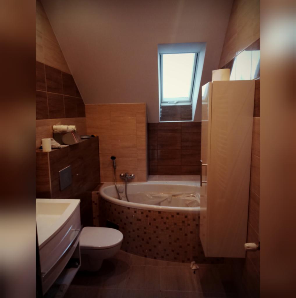 Łazienka na poddaszu w trakcie prac wykończeniowych: widoczna wanna zabezpieczona folią, częściowo ułożone płytki w odcieniach brązu, biała szafka wisząca i okno dachowe.
