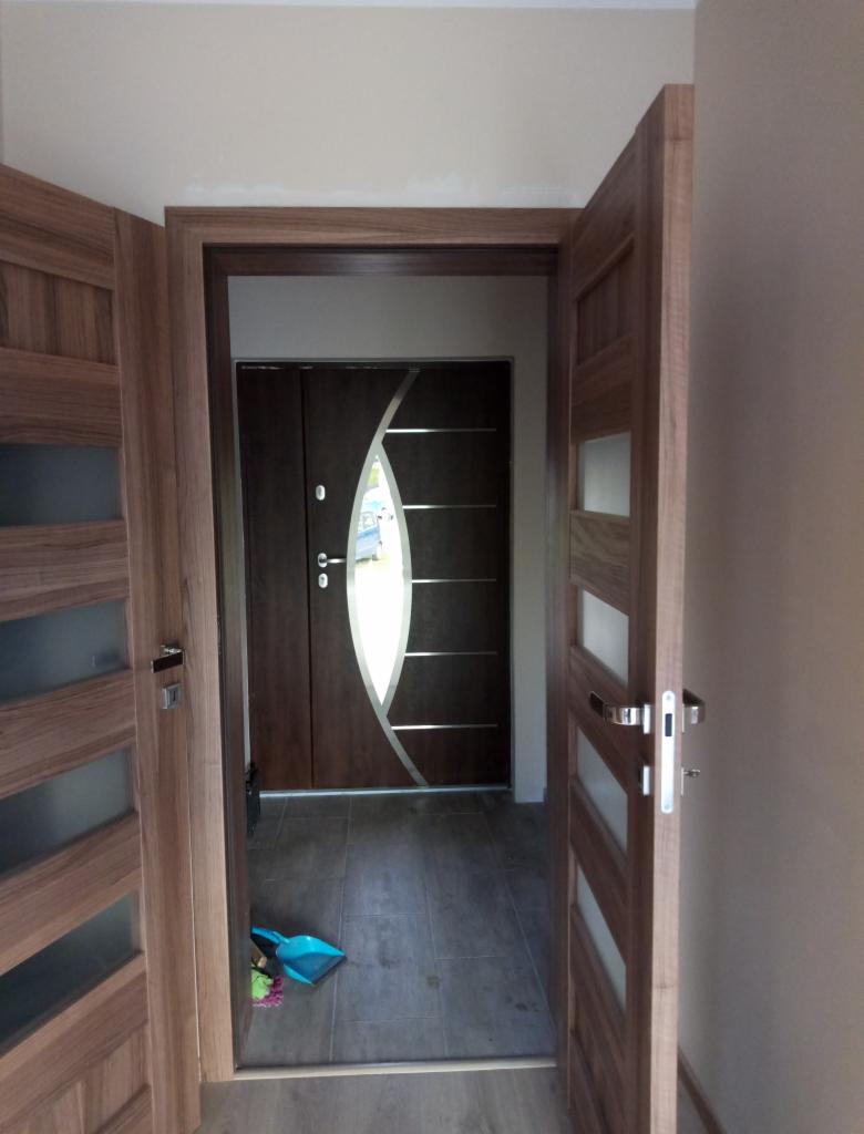 Otwarty widok na brązowe drzwi wejściowe z owalnym przeszkleniem i dekoracyjnym srebrnym paskiem, z widoczną szufelką i zmiotką na podłodze w trakcie lub po montażu.