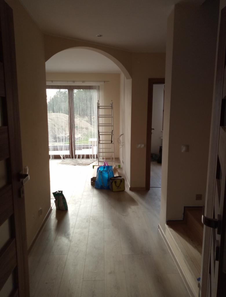 Wnętrze mieszkania w trakcie remontu: widok na korytarz z drewnianą podłogą, drabina oparta o ścianę w salonie, torby z narzędziami i materiałami budowlanymi.