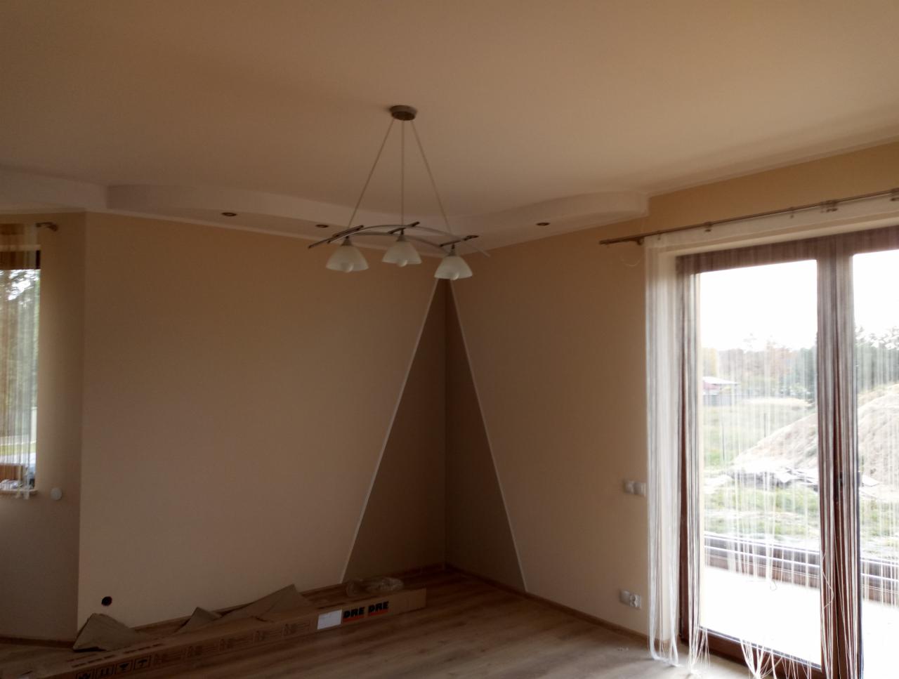 Jasne, świeżo pomalowane wnętrze pokoju z drewnianą podłogą, nowoczesną lampą sufitową i dużym oknem balkonowym z zasłonami sznurkowymi; kartony po panelach podłogowych leżą przy ścianie.