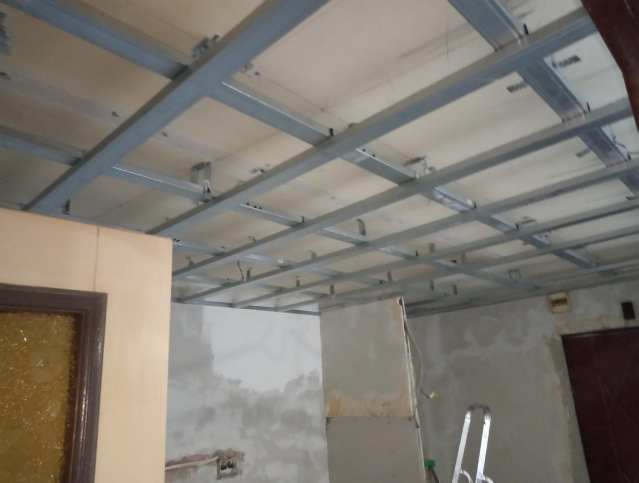 Stelaż metalowy sufitu podwieszanego z płyt kartonowo-gipsowych, widoczne profile CD i UD, surowe ściany w tle przed wykończeniem, drabina aluminiowa w pomieszczeniu.