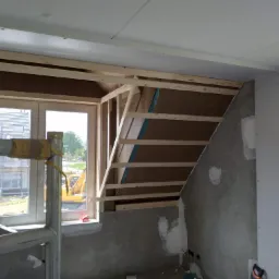 Poddasze w trakcie ocieplania: widoczne drewniane elementy konstrukcyjne, fragment izolacji, surowa ściana z widocznymi śladami gładzi, okno z widokiem na plac budowy i żółtą koparkę, fragment...