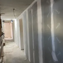 Korytarz w trakcie wykańczania z zamontowanymi płytami gipsowo-kartonowymi Habito, widoczne łączenia płyt wypełnione gipsem, surowy betonowy sufit z wystającymi przewodami elektrycznymi, w tle...