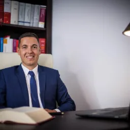 Uśmiechnięty mężczyzna w niebieskim garniturze siedzi przy biurku z otwartą książką i laptopem, za nim regał z książkami i lampka biurkowa.