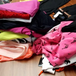 Stos pogniecionych ubrań w różnych kolorach i wzorach na drewnianym stole, w tym różowa bluza z czarnymi sylwetkami koni i żyraf, czerwona bluzka z koronką i czarna koszula z paskiem.