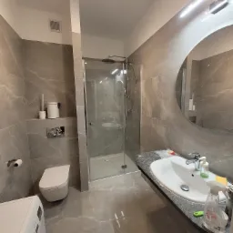 Nowoczesna łazienka w szarościach z kabiną prysznicową typu walk-in, umywalką nablatową i dużym okrągłym lustrem z podświetleniem LED, widoczna pralka z przodu.