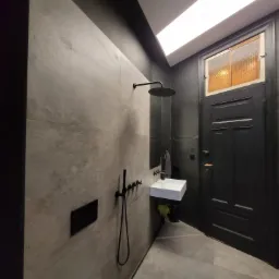 Nowoczesna łazienka w odcieniach szarości z czarnymi akcentami, deszczownicą i umywalką, oświetlona podłużną lampą sufitową, z widoczną starą, czarną, drewnianą stolarką drzwiową z pomarańczową szybą.
