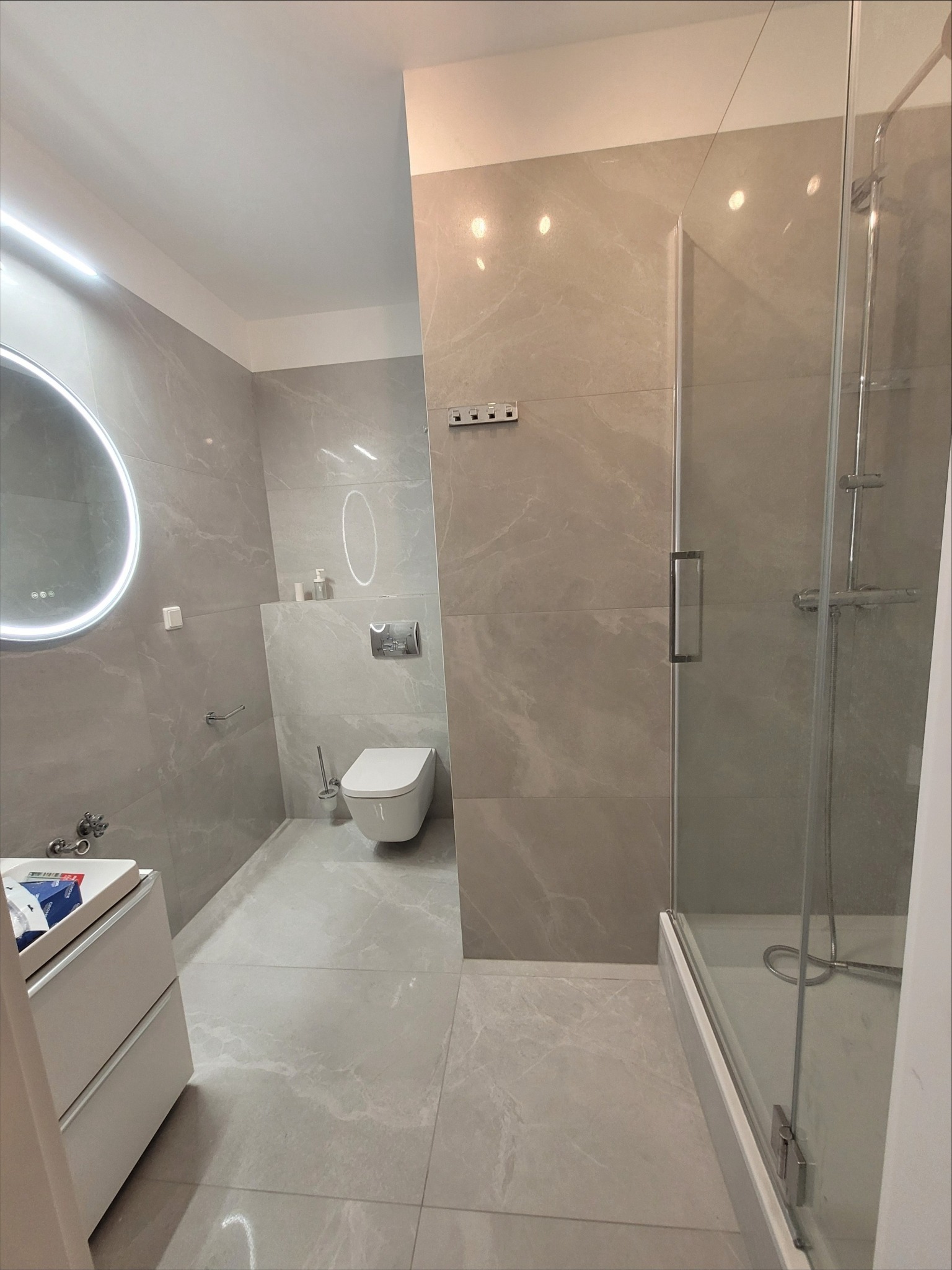 Nowoczesna łazienka z szarymi płytkami imitującymi marmur, okrągłe lustro z podświetleniem LED, biała toaleta podwieszana i kabina prysznicowa z szklanymi drzwiami.