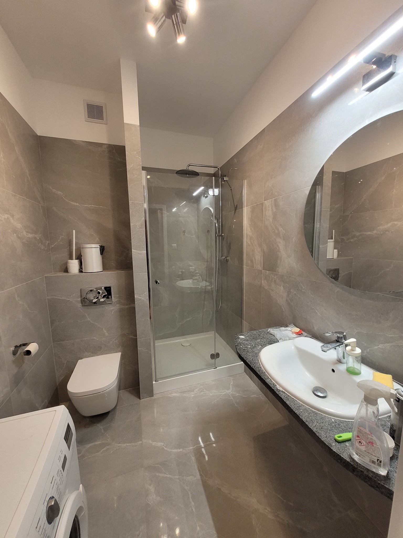 Nowoczesna łazienka w szarościach z kabiną prysznicową typu walk-in, umywalką nablatową i dużym okrągłym lustrem z podświetleniem LED, widoczna pralka z przodu.