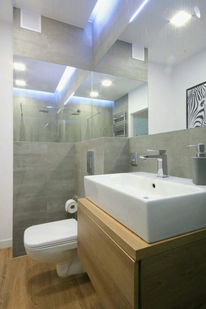 Nowoczesna łazienka z szarymi płytkami, drewnianą szafką pod umywalką i lustrem z podświetleniem LED, odbijającym kabinę prysznicową.