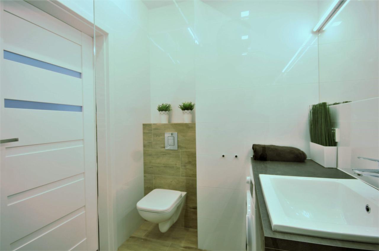 Nowoczesna łazienka z białymi płytkami, podwieszaną toaletą, białymi drzwiami z niebieskimi wstawkami i roślinami w doniczkach.