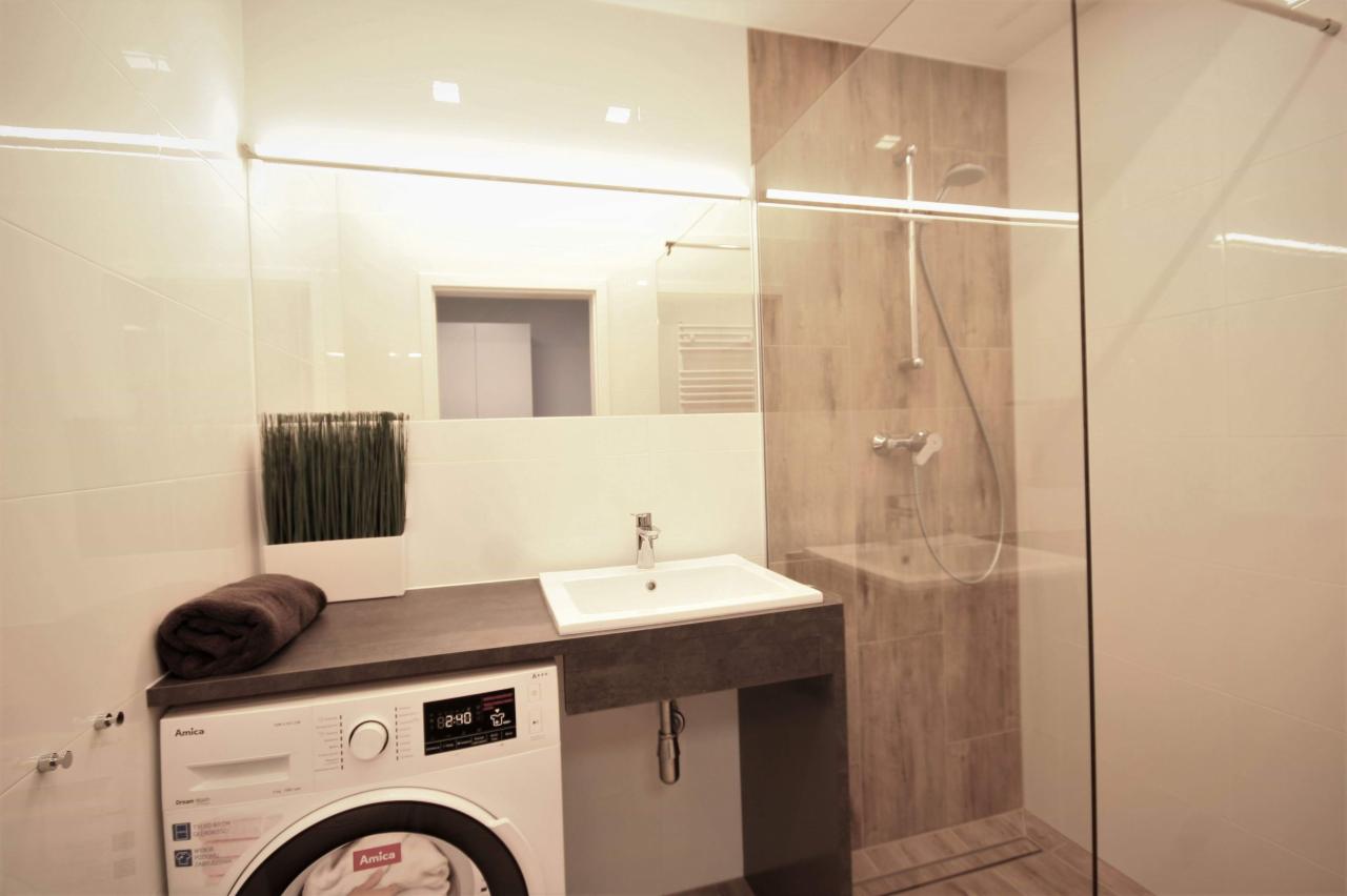 Nowoczesna łazienka z kabiną prysznicową typu walk-in, umywalką wpuszczoną w blat, pralką Amica i dużym lustrem z oświetleniem LED.