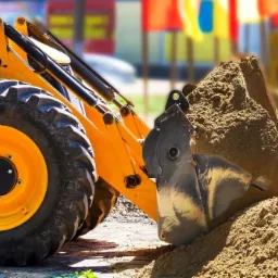 Żółta ładowarka kołowa z łyżką pełną piasku na placu budowy, w tle kolorowe flagi. Widoczne opony z wyraźnym bieżnikiem. Słońce oświetla piasek, tworząc cienie.