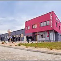 Budynek firmy z czerwonym narożnikiem i szarym dachem, widoczny z boku, z grupą osób przed wejściem i logo firmy na fasadzie.