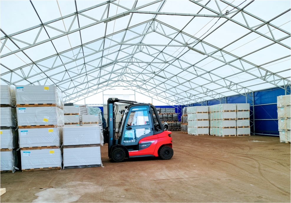 Czerwony wózek widłowy Linde w magazynie z konstrukcją stalową, obok palety z białymi opakowaniami, w tle niebieska plandeka. Transport wewnątrz magazynu.