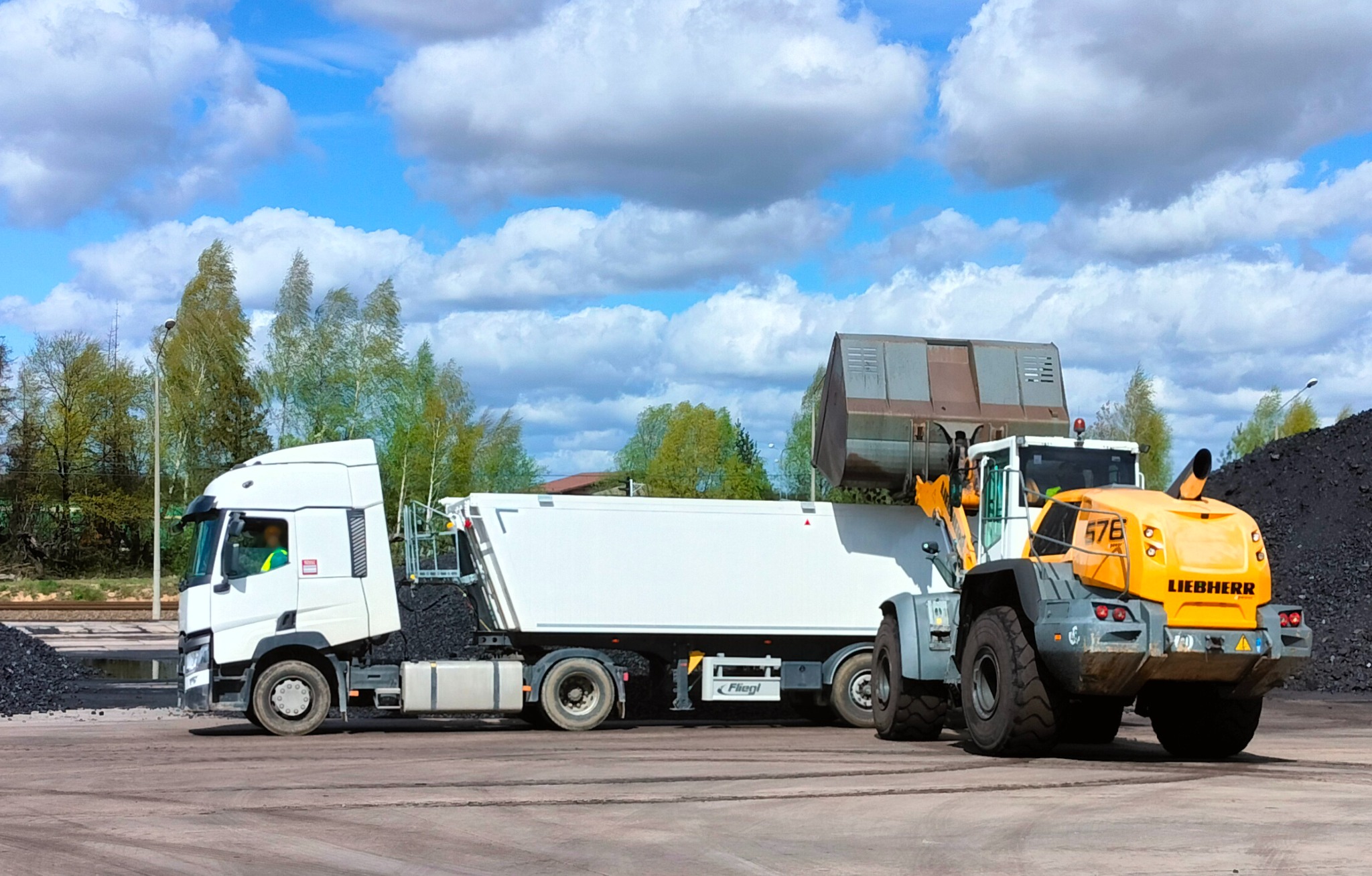 Ładowarka Liebherr nabiera węgiel do białej naczepy ciężarówki na tle błękitnego nieba z chmurami, widok z boku, na ziemi węgiel.