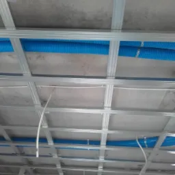 Metalowa konstrukcja sufitu podwieszanego z widocznymi niebieskimi rurami wentylacyjnymi, zamontowanymi nad płytami kartonowo-gipsowymi, widok z dołu.