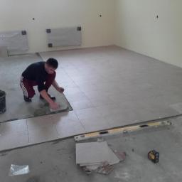 Tomix Tomasz Wysiadły - Pracownik w trakcie układania jasnych, kwadratowych płytek podłogowych w dużym pomieszczeniu, z widocznym wiadrem z zaprawą, poziomicą i laserowym miernikiem odległości.