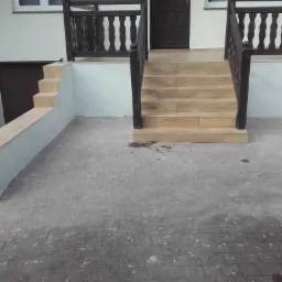 Schody wejściowe wyłożone płytkami imitującymi drewno, z ciemnymi balustradami, prowadzące do ciemnych drzwi wejściowych, z widoczną kostką brukową na podjeździe i fragmentem elewacji budynku.