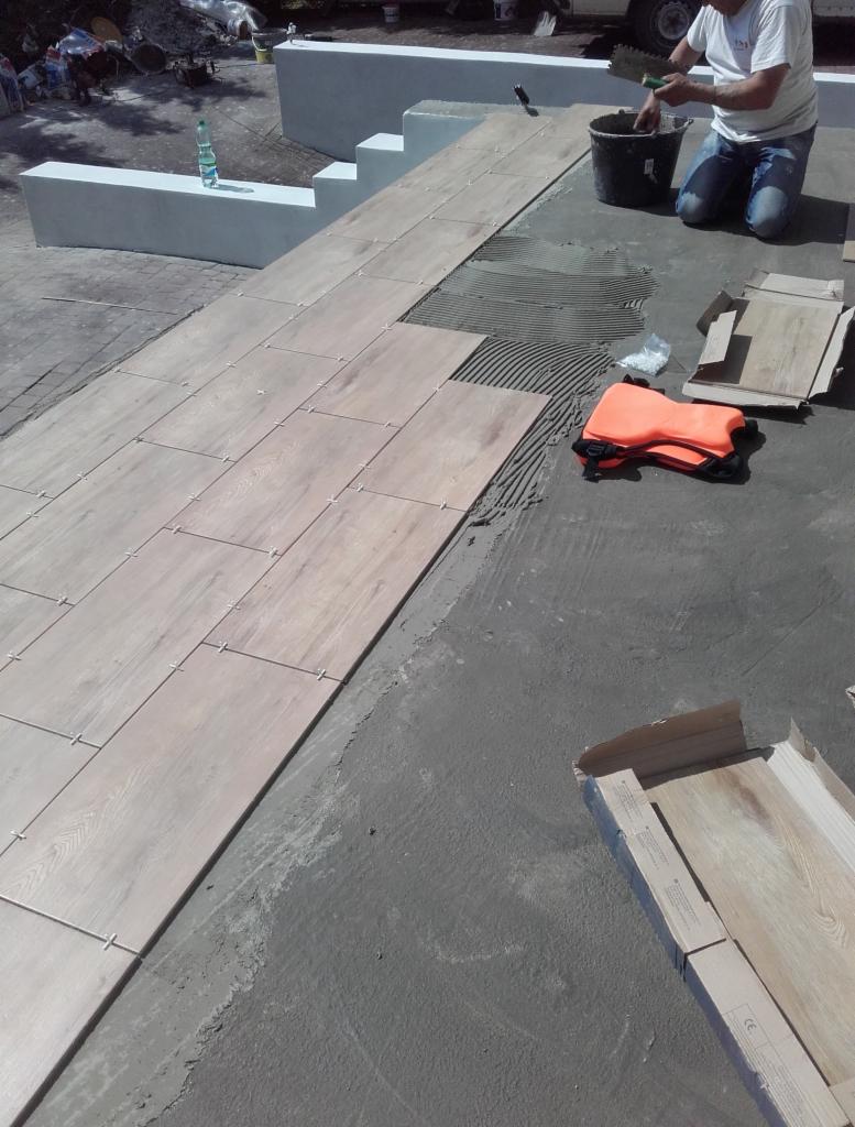 Układanie podłużnych płytek imitujących drewno na zewnątrz budynku, widoczne krzyżyki dystansowe, pracownik nakłada klej z wiadra za pomocą pacy zębatej, obok leżą puste kartony po płytkach...