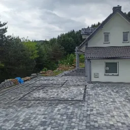 Szara kostka brukowa ułożona na podjeździe przed domem z jasną elewacją, z widocznymi paletami kostki i taczką w tle, na zboczu zalesionego wzgórza.