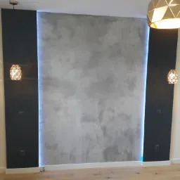 Szara ściana dekoracyjna z efektem betonu, oświetlona pionowymi pasami LED, flankowana czarnymi panelami, podwieszone lampy z ażurowymi kloszami, jasna drewniana podłoga.