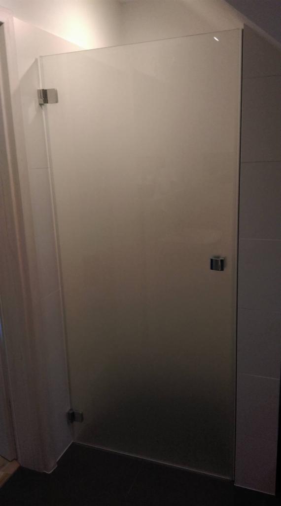 Szklana ścianka prysznicowa z chromowanymi zawiasami, widok z boku, ciemna podłoga i jasne płytki na ścianie.