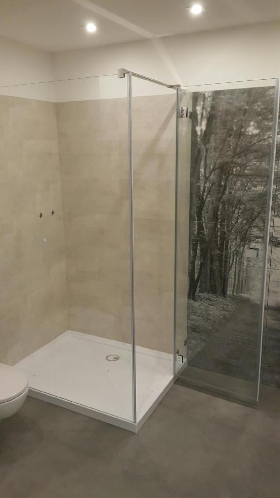 Nowoczesna kabina prysznicowa z szklaną ścianką z motywem leśnym, biały brodzik i częściowo widoczna toaleta w łazience wykończonej beżowymi płytkami.
